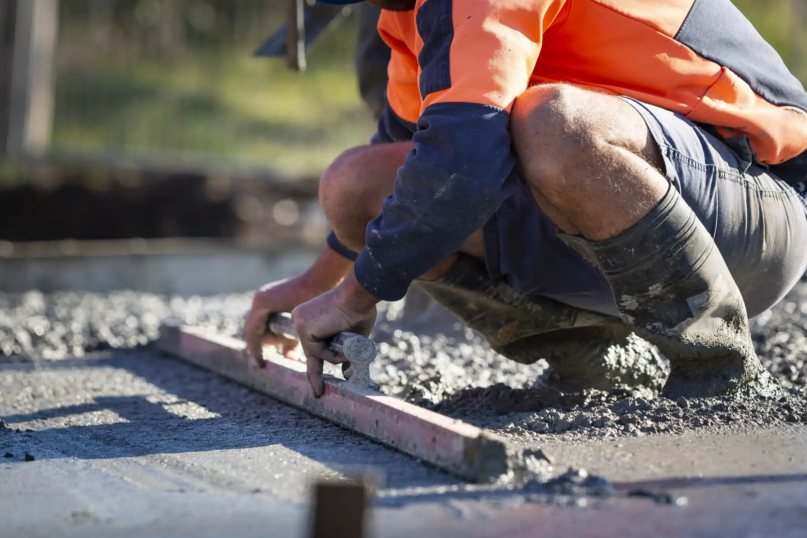 On a sunny outdoor site, a construction worker in an orange and blue uniform expertly levels wet concrete, demonstrating advanced hardscaping skills. Kneeling in rubber boots, the worker concentrates on perfecting the surface with precision.