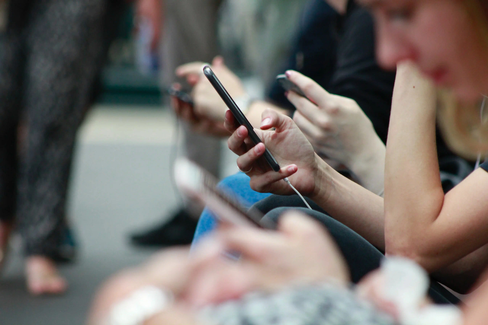 A group of individuals sits closely, each intently focused on their smartphones, engaging with the newest Los Angeles SEO trends. Firmly holding their devices, white earphone cords are clearly visible. The blurred foreground and background underscore the primary focus on the phones and hands.