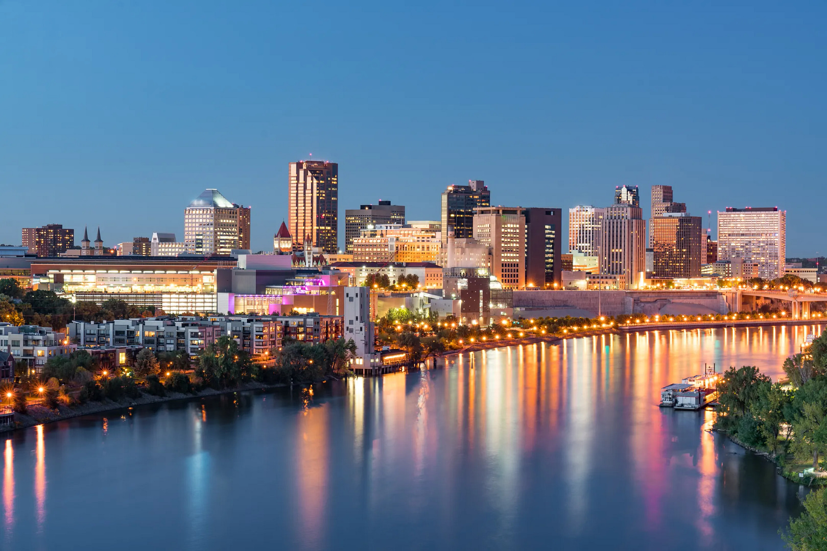 Experience the tranquil beauty of an illuminated city skyline reflected on a calm river. Modern skyscrapers elegantly blend with historic architecture, echoing St. Paul's charm. Lush riverbank vegetation complements a clear, deep blue sky, creating a serene urban masterpiece.