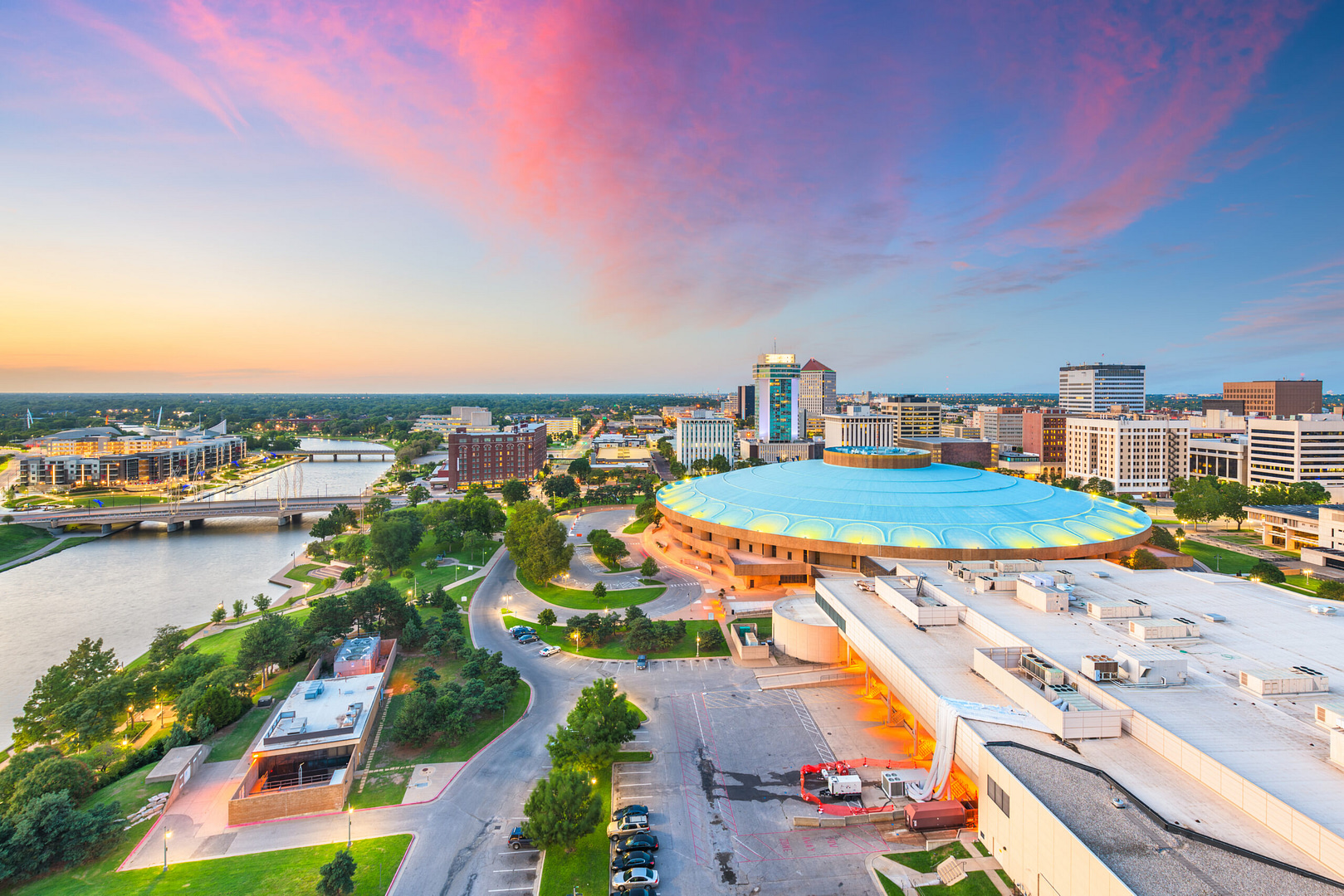 Discover Wichita's unique harmony of nature and architecture with an aerial view at sunset. A prominent circular building with a blue-green roof stands near the river, framed by city structures and a bridge. The picturesque scene of vibrant pink clouds mirrors Wichita’s evolving SEO landscape.