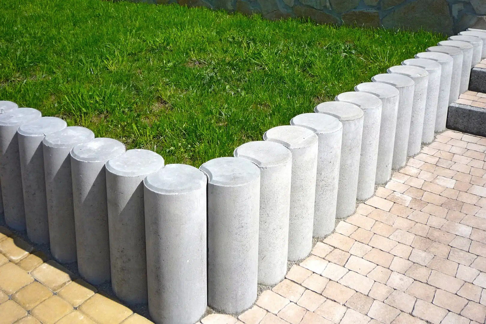 Enhance your landscape with a striking design: cylindrical concrete posts frame lush green grass, seamlessly connecting light-colored pavers to a yellow-tiled walkway. This showcases exceptional landscaping prowess, reflective of top-tier Landscaping PPC strategies.