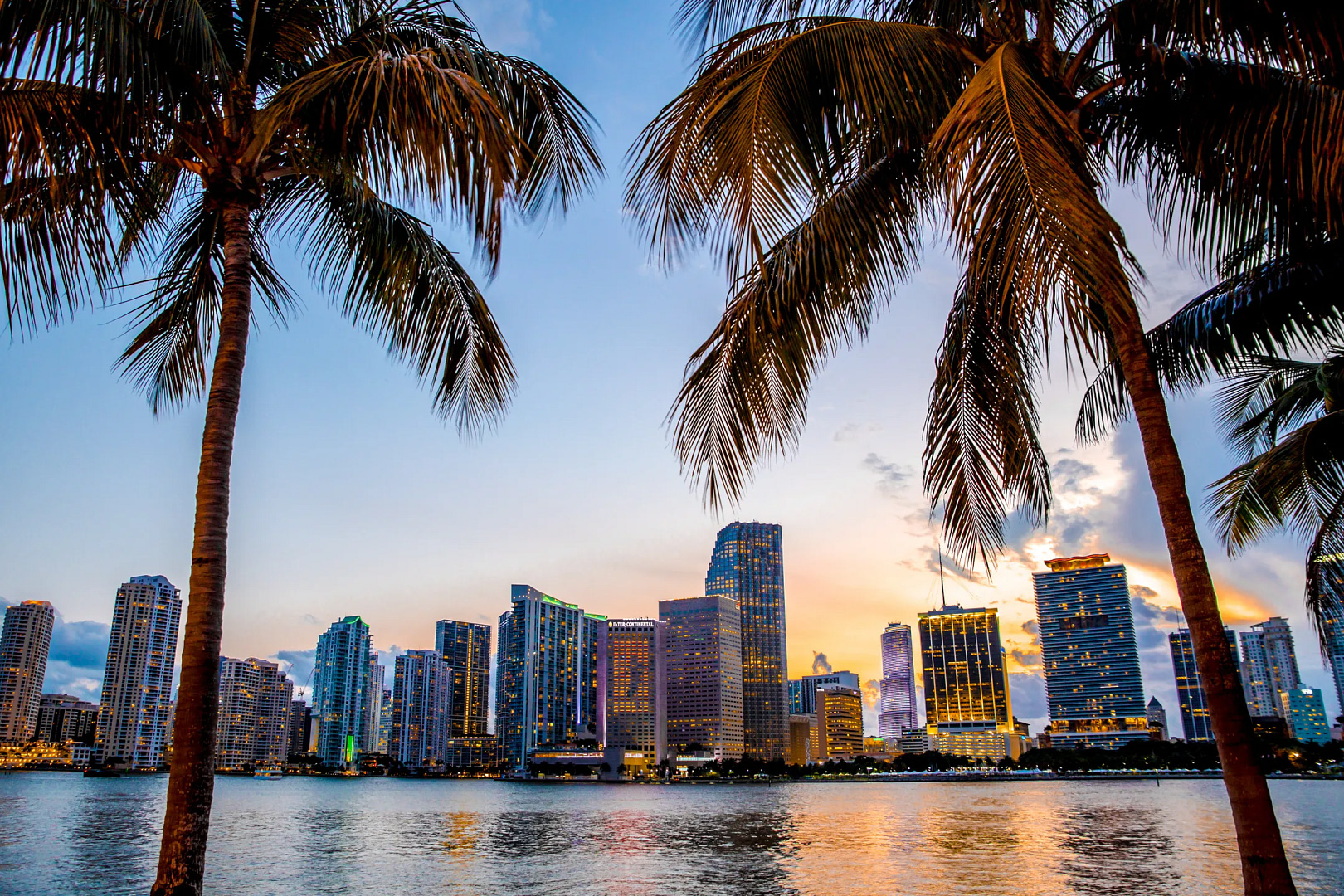 Experience the allure of Miami with a sunset city skyline featuring sleek skyscrapers mirrored in tranquil waters. Framed by palm trees, this tropical scene radiates the vibrant hues of orange, pink, and blue, perfectly embodying inspiring Miami web design aesthetics.