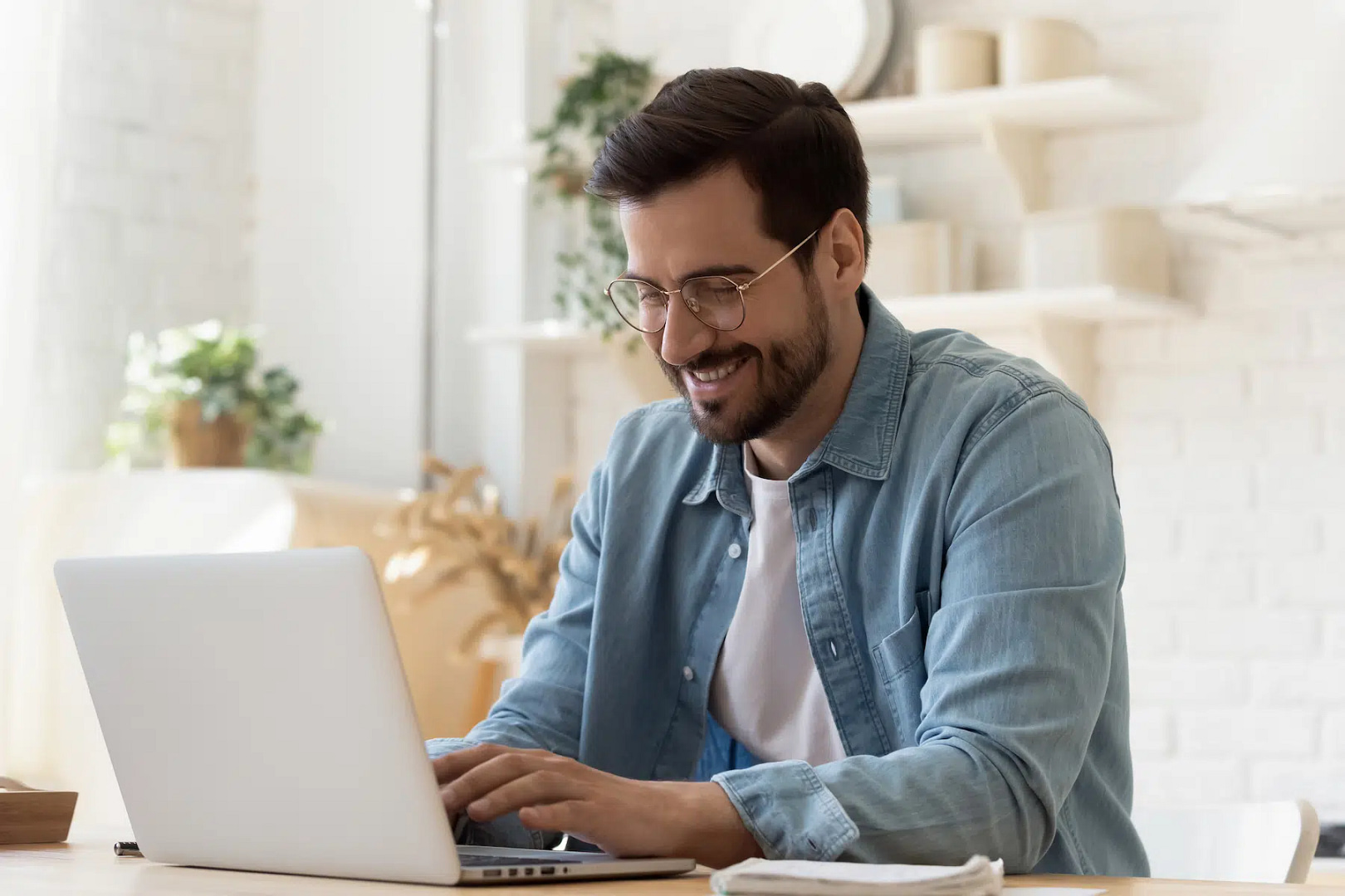 A bearded man wearing glasses is intently focused on Milwaukee web design projects at his laptop in a well-lit room, with plants and shelves enhancing the inviting ambiance.