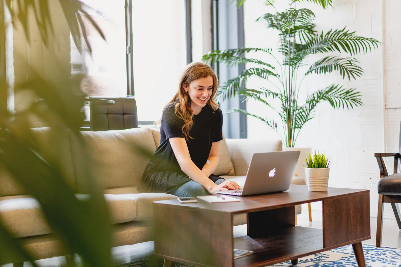 A woman sits on a beige sofa, intently researching San Diego SEO strategies on her laptop. Natural light floods the room through large windows, highlighting green plants and a patterned rug.