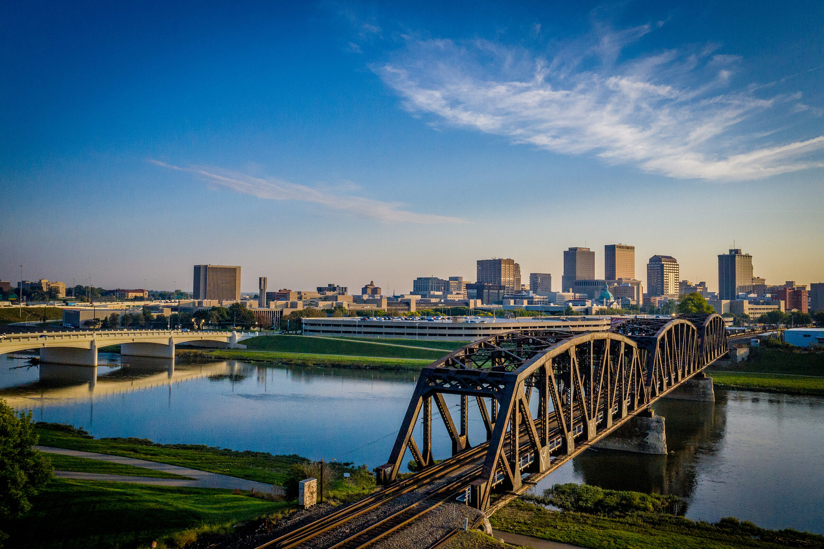 Experience the enchanting aerial view of a cityscape at sunset, featuring a large river spanned by two parallel bridges. The skyline, echoing Dayton's charm, rises majestically beneath a clear blue sky adorned with delicate wispy clouds.
