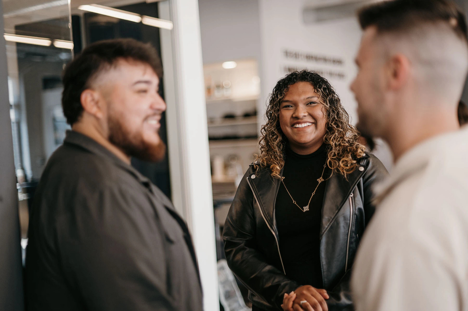 Three individuals are engaged in an indoor conversation. A person with curly hair and a black leather jacket smiles at two others: one has a beard and wears a dark jacket, while the other has short hair and sports a light shirt. The modern environment, featuring visible shelves and decor, underscores their focus on career opportunities.