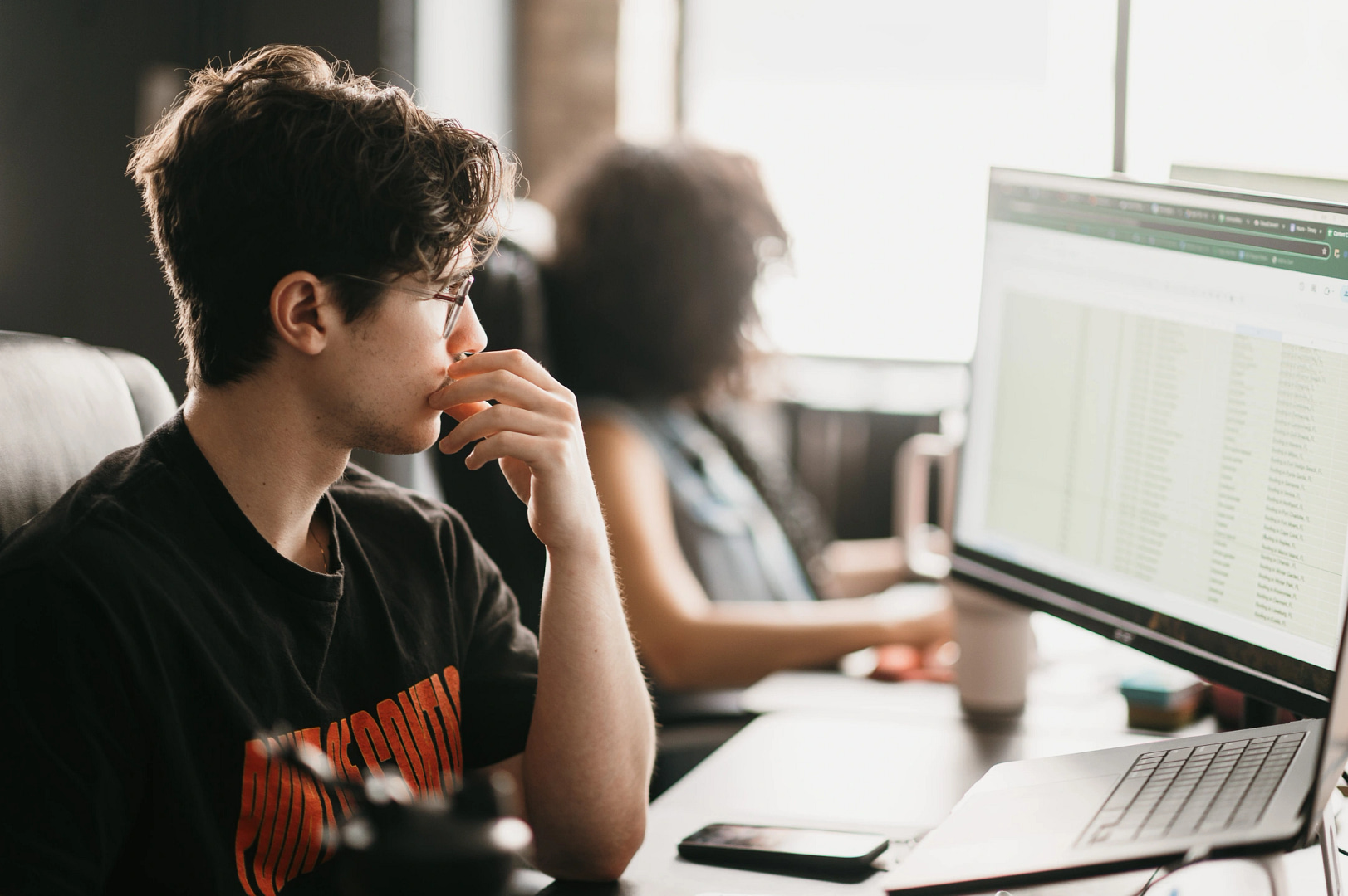 A person in glasses and a black "UNKNOWN" t-shirt intently examines a Minnesota SEO strategy spreadsheet at their desk, while another individual works on their computer in the background.