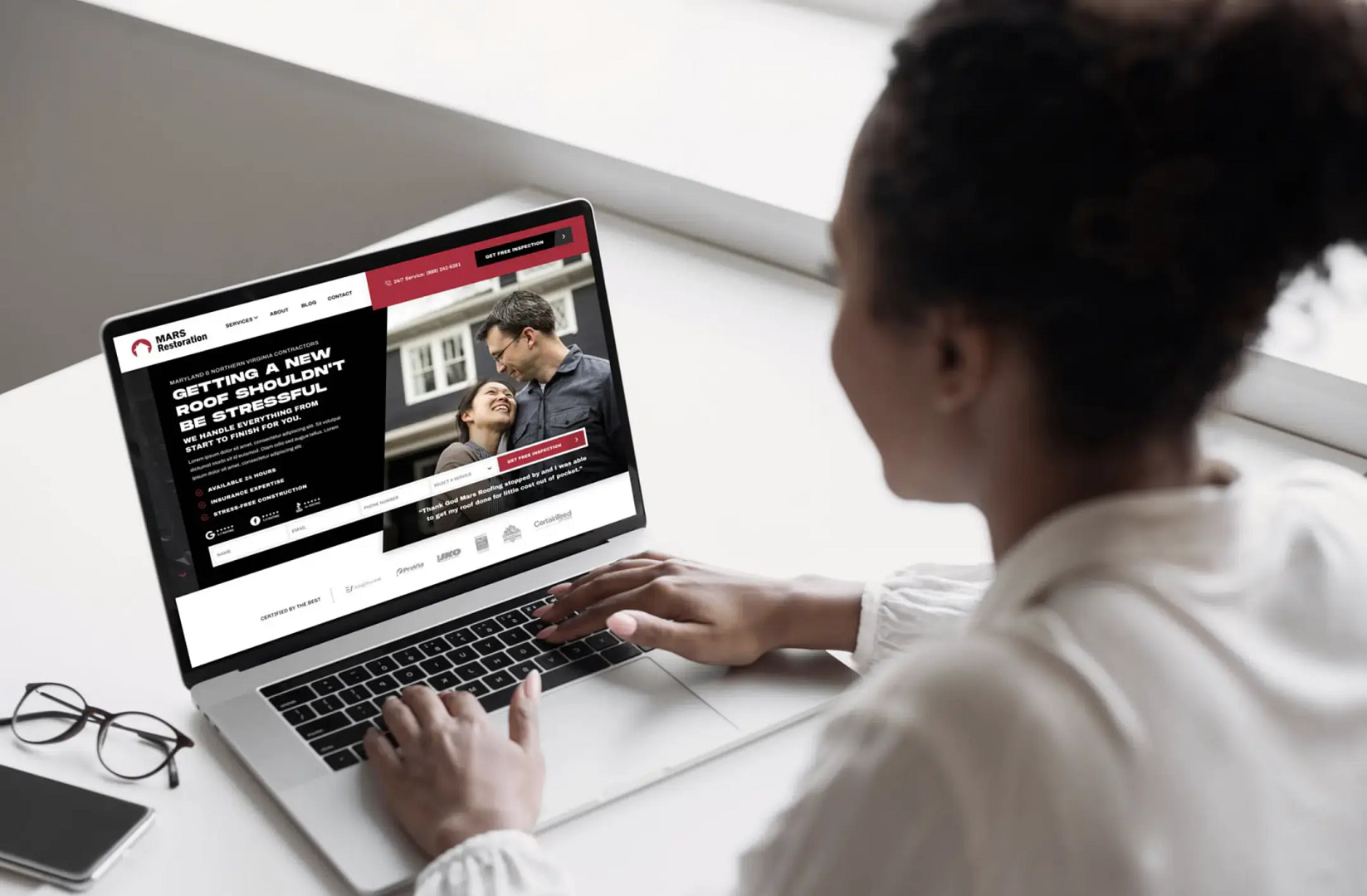 An individual dressed in a white top is seated at a bright desk, browsing a roofing services website on their laptop. The site, crafted with the refined touch of Madison web design, prominently features an image of a couple sharing a glance, accompanied by text promoting stress-free solutions.