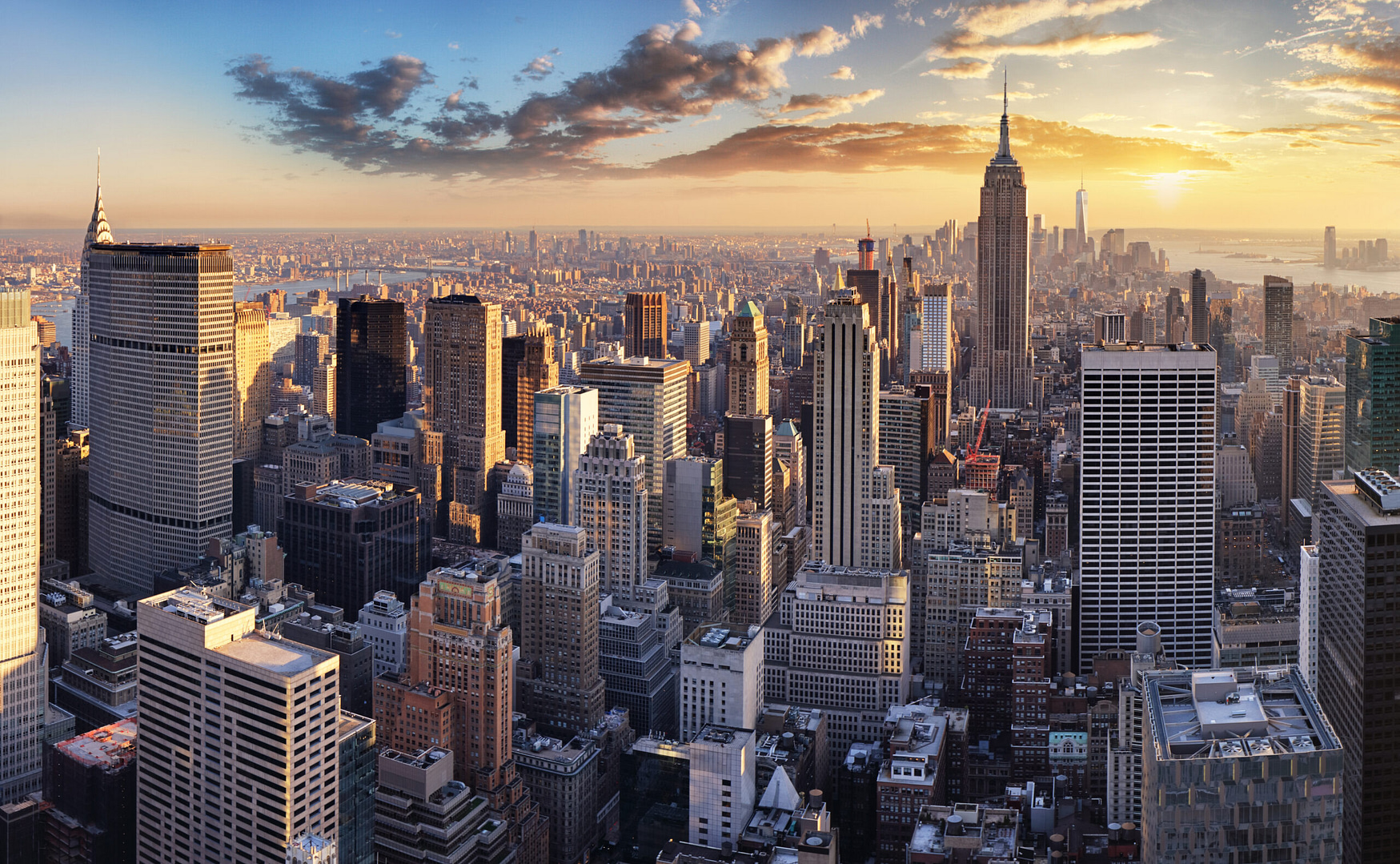 Experience the iconic New York skyline at sunset, where towering skyscrapers and a distinctive pointed building are illuminated in a warm glow. The river sparkles under a partially cloudy sky, embodying the city's captivating spirit and inspiration.