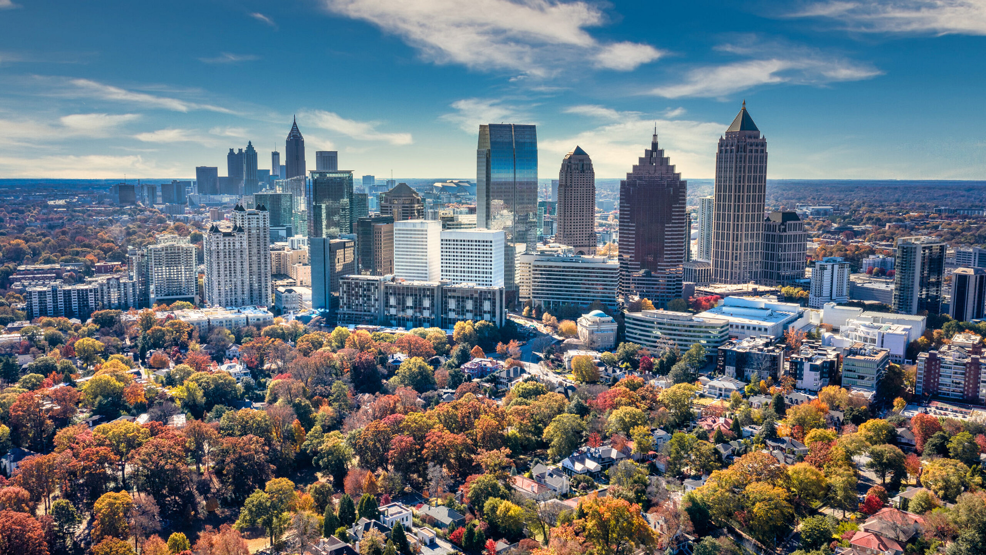Experience Atlanta's skyline through a breathtaking aerial view of modern skyscrapers set against a partly cloudy sky. In the foreground, a vibrant park displays autumn’s finest colors—green, fiery reds, and oranges—perfectly capturing the city's harmony of urban life and natural beauty.