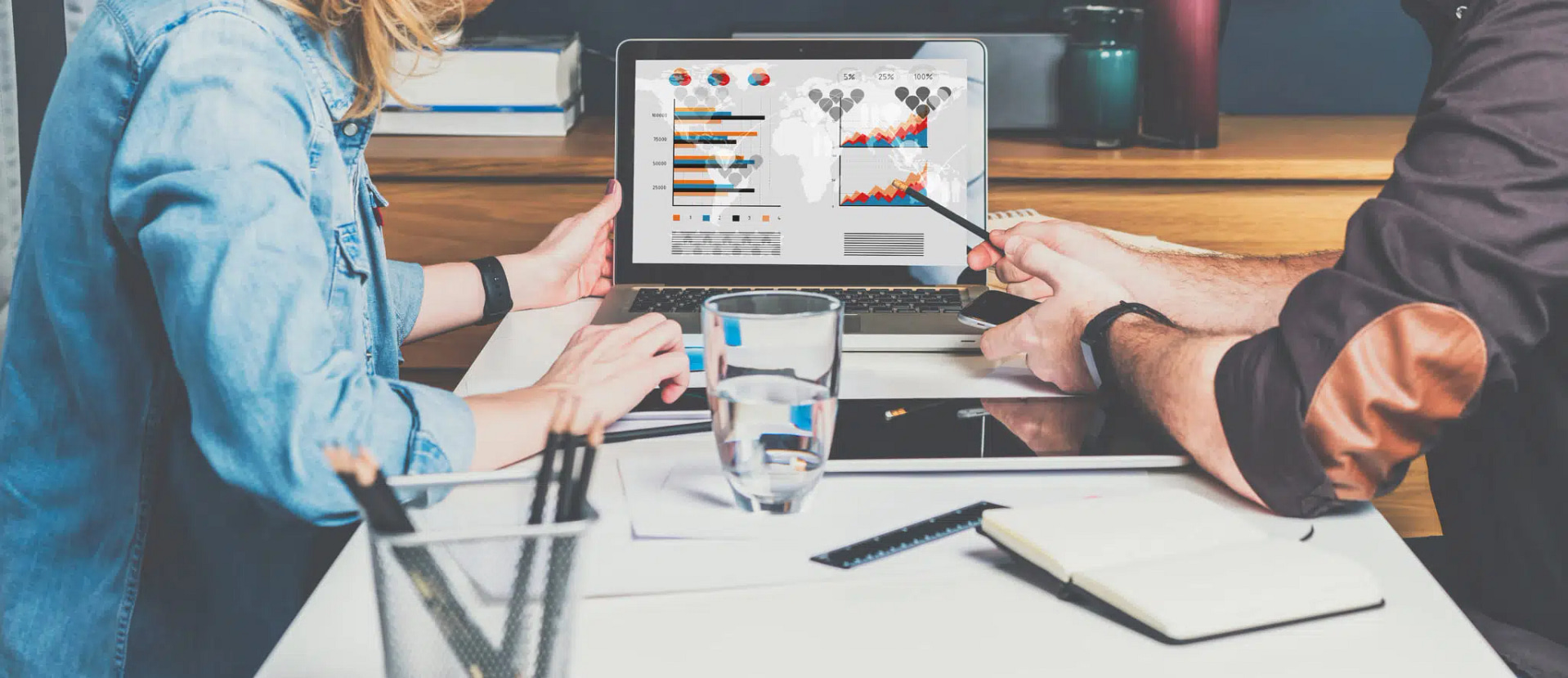 Two individuals sit at a table reviewing graphs on a laptop, surrounded by a glass of water, a notebook, a smartphone, and pencils. One points to the screen, discussing ways Lincoln SEO strategies could improve the outcomes shown in the colorful charts.