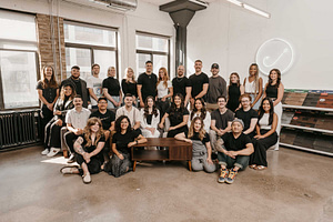 A team of 23 stands confidently before large windows, symbolizing the core of home services marketing. While most stand, some are seated near a wooden bench, framed by sample shelves and highlighted with modern neon lighting.