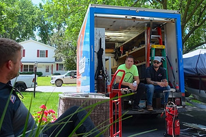 Nick Thelen (owner of HVAC and Plumbing company Thelen Mechanical) and Tim Brown in a lime green Hook Agency shirt sit inside the back of an open plumbing and hvac truck, showcasing Hook Agency's Process and really getting to know it's clients industries more deeply, on a residential street. Another person sits outside the truck surrounded by trees and houses, a Roofer Josh Swisher of Northface Construction who has been working with Hook Agency for 9 years. They chat warmly and discuss business and Hook Agencies Process. Various tools and equipment are visible inside, illustrating a systematic organization, because Hook Agency works with the best home service businesses.