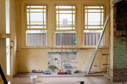 In the midst of renovation, a room with peeling paint, exposed brick, and three expansive windows signals an active home remodeling project. An upright broom leans against the wall beside a floor scattered with tools. Sunlight streams through sheer blinds, casting a warm glow on this soon-to-be showcase for Hook Agency's remodeling expertise.