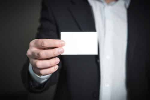 A person in a black suit presents a blank white business card to the camera, poised to make an impact at the upcoming home improvement conference.
