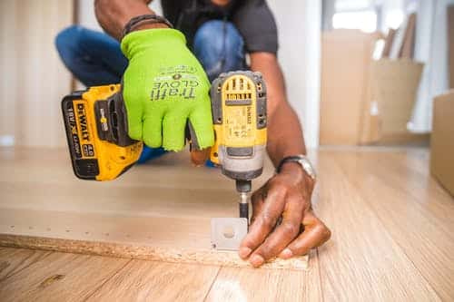 On our website, you'll find this image: A person kneels on a wooden floor, expertly using a yellow cordless drill to install a screw into a wooden plank. One green-gloved hand grips the drill while the other steadies the task. The backdrop is a bright room with softly blurred furniture, embodying the essence of home improvement marketing.