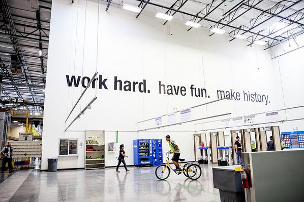 Experience the dynamic energy of our industrial office space at Hook Agency, boasting high ceilings and prominently displaying Amazon's inspiring slogan: "Work Hard. Have Fun. Make History." Observe the bustling activity, with individuals engaging collaboratively and one riding a yellow bike through the vibrant workspace.