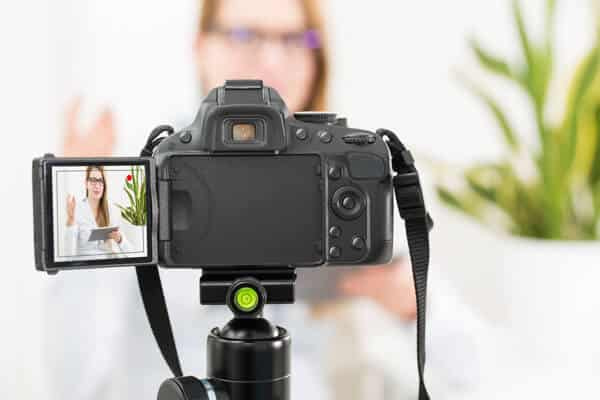 A camera on a tripod captures a presenter discussing video marketing strategies indoors, with a plant nearby. The person is holding a tablet and appears on the camera's screen against a blurred background.