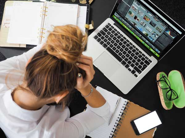 A person sits at a desk with their head in their hands, surrounded by chaos. An open laptop displays an image editing program, hinting at the struggle of avoiding SEO pitfalls. Notebooks, a smartphone, and scattered papers reveal the complex world of navigating common SEO errors.