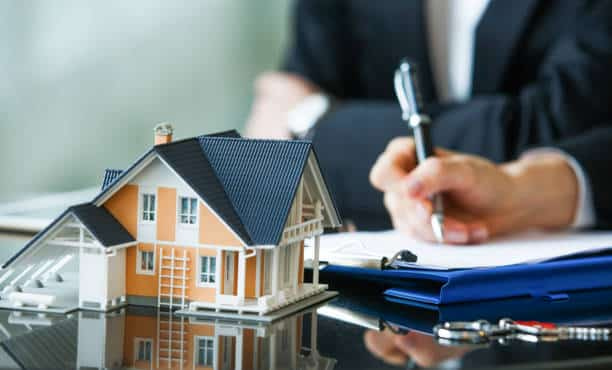 On a desk, a small house model is prominently displayed beside a set of keys, while an individual in a suit meticulously signs documents on a clipboard in the background. This scene underscores the complexities involved in roof sales commissions within real estate transactions.