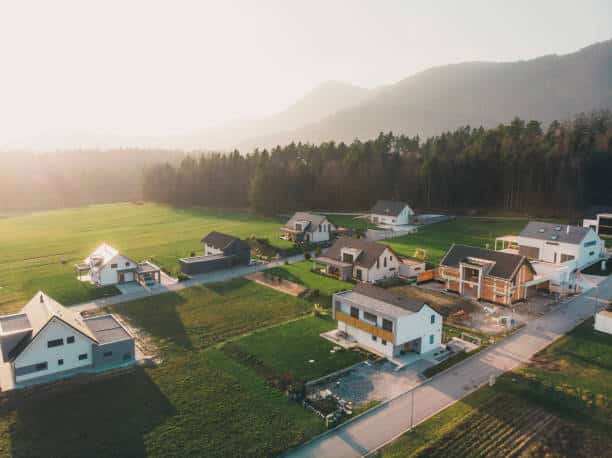 Experience the success of Dan Walrack's roofing sales through an aerial view of a suburban neighborhood. Modern homes with expertly crafted roofs are nestled among green fields, trees, and distant hills. The warm glow of early morning sunlight highlights the enhanced curb appeal these roofs provide.