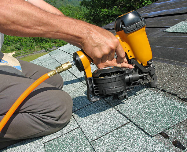 An expert uses a top-rated yellow pneumatic nail gun for 2024 to affix green and gray asphalt shingles on a roof. The tool is connected via an orange air hose, set against a backdrop of lush greenery.