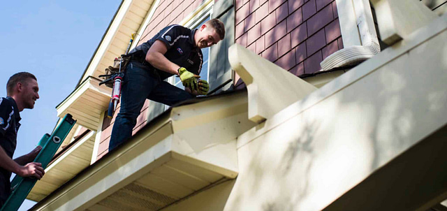 Two workers expertly install a solar panel on a beige-sided house with maroon shingles. One secures the panel from the roof, while the other efficiently passes tools from a ladder below.