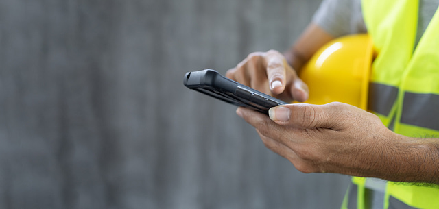 A person in a safety vest holds a yellow hard hat under one arm and checks their smartphone—potentially searching for home services—set against a blurred gray background.