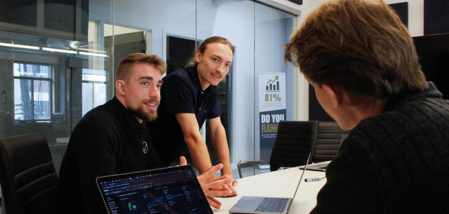 In a business meeting room, two individuals stand near a laptop sharing SEO tips with a third person who is seated and engaged. Behind them, a glass window displays an impactful 81% statistic poster.