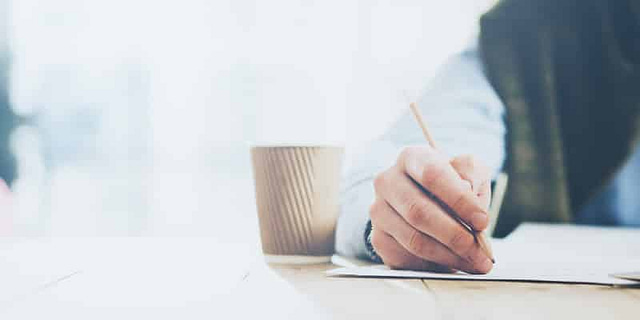 A close-up of an intern's hand writing with a pencil on a piece of paper captures the essence of content production. A disposable coffee cup with a lid sits beside the hand. The softly blurred background lends a bright, airy feel to the scene, emphasizing the casual tabletop setting and focus on creativity.