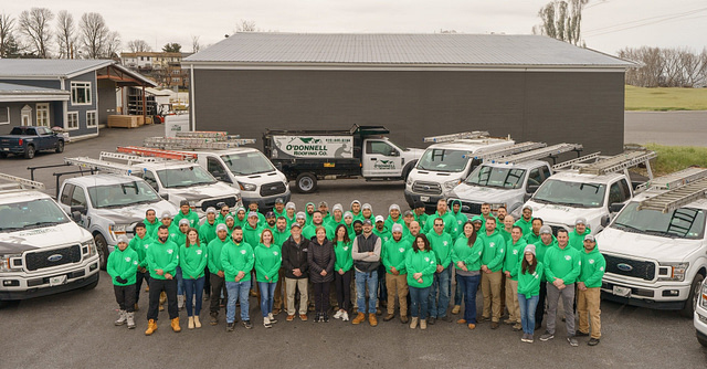 A unified team—representing the ideal Hook Agency client—poses for a group photo in front of multiple white utility trucks and a black O'Donnell Roofing Co. truck, outside a gray commercial building. Their matching green shirts reflect professionalism and strong company branding.