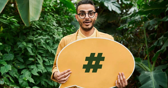 In a vibrant garden setting, an individual with glasses enthusiastically holds a large yellow speech bubble showcasing a hashtag symbol, illustrating the power of social signals in boosting SEO rankings. Dressed in a beige jacket, they exude excitement at this digital revelation.