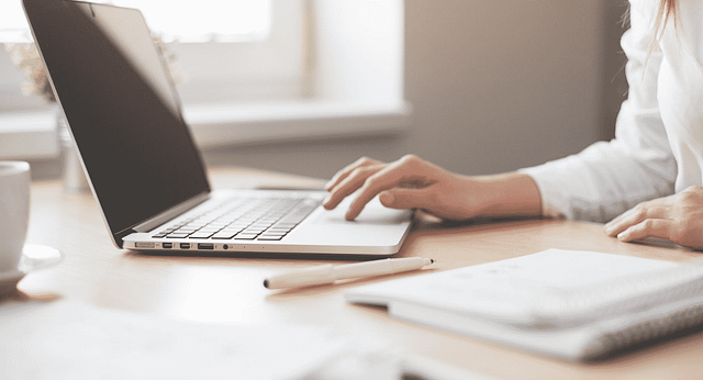 A person, likely a marketing director, types on a laptop at a desk scattered with essential marketing tools like notebooks and a pen. Sunlight filters through a window, casting a warm glow on the organized workspace. The person wears a white shirt in this well-lit setting.