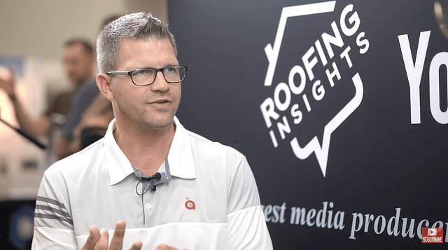 A man in a white polo shirt passionately explains strategies for roofing companies to overcome bottlenecks. The "ROOFING INSIGHTS" backdrop is prominently displayed, with people and equipment softly blurred in the background.