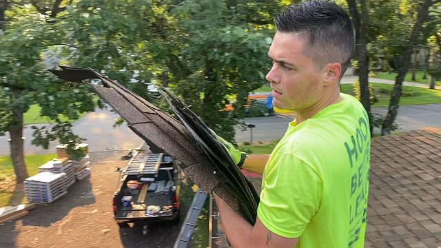 A roofing professional in a bright yellow shirt stands on a roof, confidently holding several shingles—demonstrating our deep understanding of the roofing industry. In the background, trees and a work truck loaded with roofing materials reinforce our firsthand knowledge of your business.