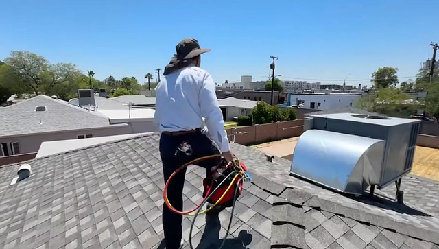 On the Hook Agency website: A uniformed professional, donned in a hat, is positioned atop a shingle-covered rooftop, firmly grasping HVAC equipment. Surrounding homes and trees extend beneath a pristine blue sky characteristic of 2025.