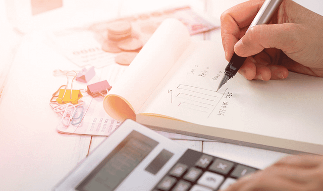 A person writing on a notepad with a pen, next to a calculator, breaks down the marketing budget. The table is cluttered with paper, clips, and coins, suggesting a financial theme involving calculations or budgeting.