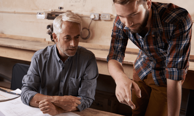 In a workshop setting, two men collaborate over a blueprint on a wooden table. One, seated with gray hair in a blue shirt, and the other standing with a beard in a plaid shirt, exemplify the teamwork among contractors focused on increasing their project's sales closing rate.