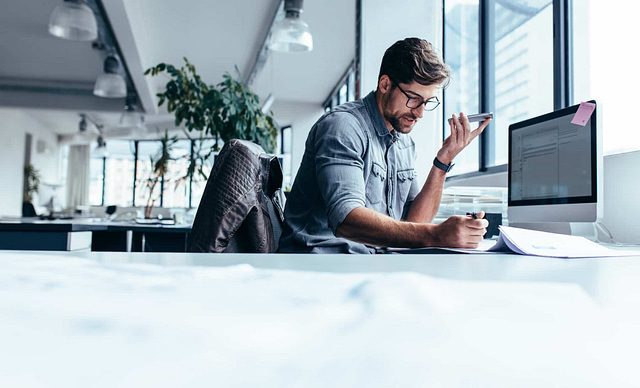 In a modern office, a professional focuses on niche provider strategies, speaking into his smartphone with pen in hand. He is deeply engaged in keyword research at his cluttered desk, illuminated by large windows and a jacket casually draped over his chair.