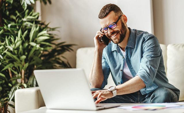 A man in glasses and a denim shirt sits on a couch, smiling as he speaks on his smartphone and types on his laptop. He's likely discussing an SEO strategy. A leafy plant enhances the tranquil setting.