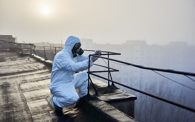 On Hook Agency's website, an individual dressed in a white hazmat suit and gas mask crouches on a foggy rooftop, gripping the railing wires. The backdrop features a mist-covered urban skyline with buildings shrouded in haze and an eerie glow from the dim sun.