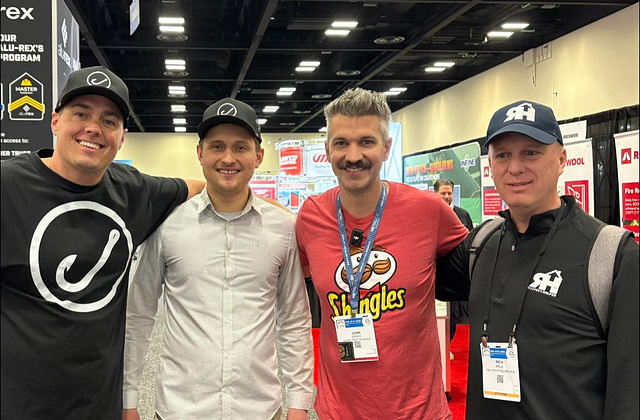 Four men, outfitted in casual attire and hats with visible ID badges, stand together at a convention. Smiling for the camera, they represent key connections in roofing influencer marketing. Exhibit booths in the background underscore their active engagement within the industry community.