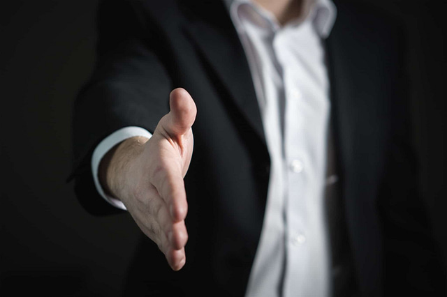 A person in a dark suit confidently extends their right hand for a handshake against a shadowy backdrop, breaking stereotypes about salespeople with an authentic gesture of welcome or agreement.