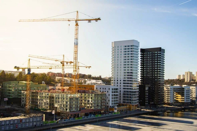 A vibrant cityscape with cranes and high-rises under clear skies showcases the dynamic pace of urban development. Modern buildings rise near waterfronts, perfectly capturing construction companies in action, all bathed in warm sunlight.