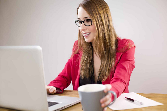 A professional woman in a red blazer, wearing glasses, confidently smiles as she works on her laptop. Holding a cup in her right hand, she is ready to seize new opportunities. A pen and notepad rest on the wooden table beside her, set against a plain light background that emphasizes focus and professionalism.
