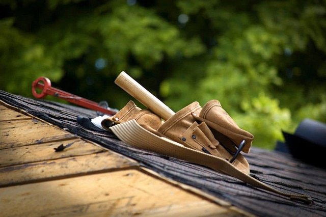 A tool belt equipped with a hammer, wrench, and other essentials lies on a rooftop under construction, symbolizing the expertise of roofing contractors. In the background, blurred green foliage offers a tranquil contrast to this dynamic work environment.
