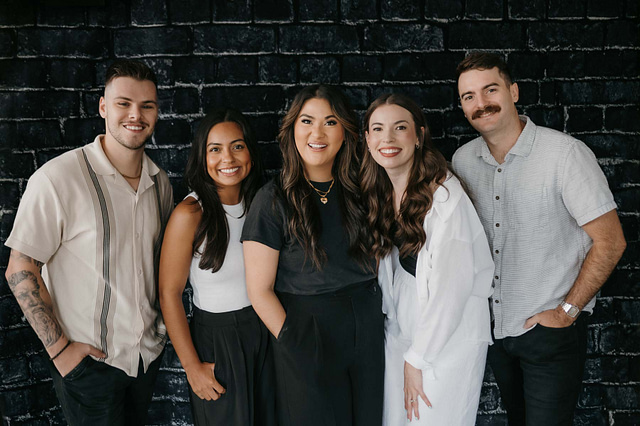 A group of five people vinny claudia, maggie, Sydnee, and skyler, embodying the spirit of camaraderie and giving a glimpse of what to expect in genuine friendships, stands close together, smiling at the camera. Positioned against a dark brick wall, the group features two men and three women, all dressed in casual attire.