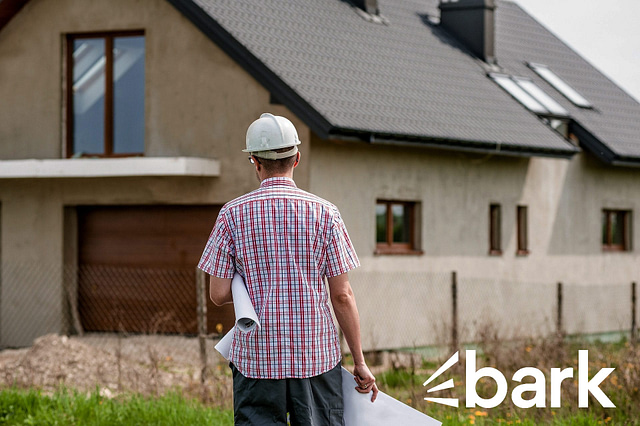 Man in a white hard hat and plaid shirt holding blueprints stands in front of an unfinished two-story house with a sloped roof. Raw concrete is visible, and a wire fence surrounds the property. "Bark Reviews" appears in the lower right corner, suggesting insights into home services and contractors.
