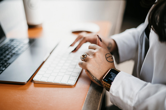 A tattooed hand with rings and a smartwatch types on a white keyboard. Nearby on the orange desk are a calculator and, in the background, a blurred coffee cup.