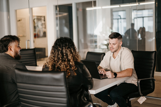 Three marketing professionals meet at a conference table in a modern office. One types on a laptop as the others engage and listen. Glass walls and office chairs define the contemporary workspace—capturing Hook Agency’s collaborative approach in action.